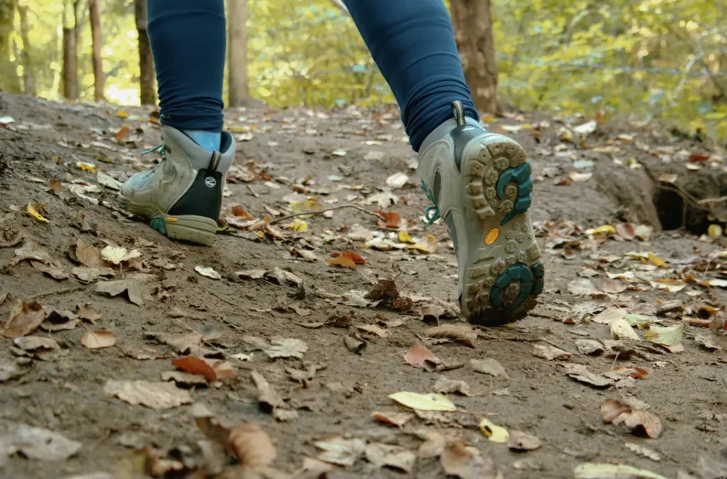 Low-angle shot of grey minimalist trail shoes with Vibram sole stepping onto a dirt path in an autumn forest, right foot mid-push-off with sole visible, illustrating heel breathing hiking in motion