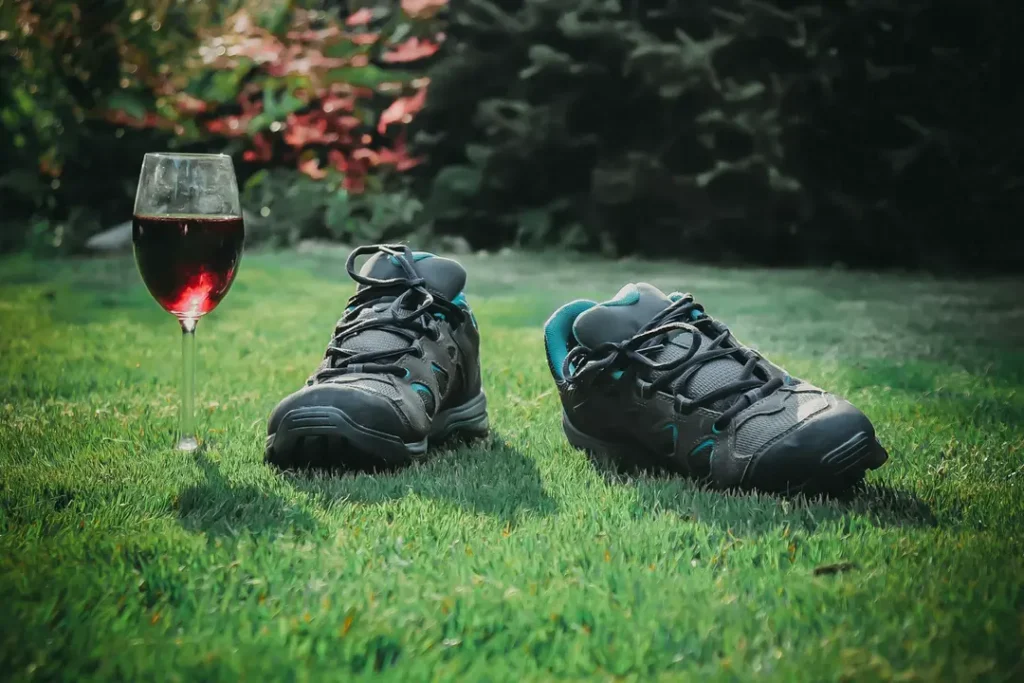 A pair of minimalist trail shoes resting on green grass beside a glass of red wine in natural afternoon light, evoking the quiet reward after a walk and the sensory return home that follows every Sensory Roaming practice