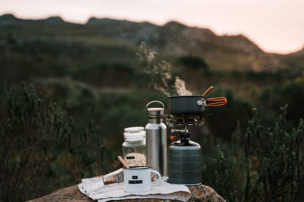 A small titanium cup steaming beside a compact canister stove on a flat rock at dusk, a single wooden spoon in the cup, mountains blurred behind in fading light, illustrating minimalist backcountry cooking