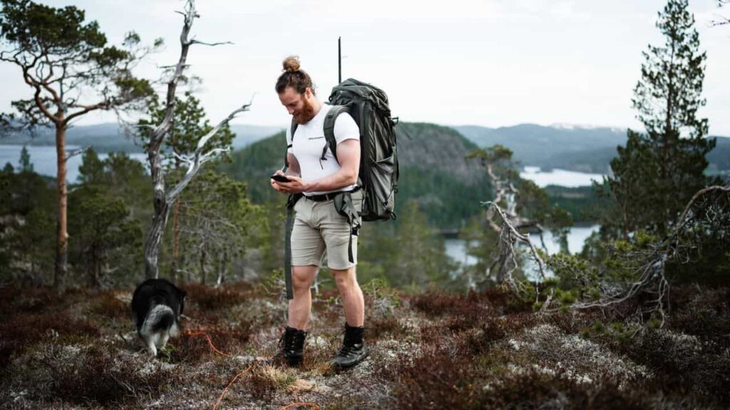 A hiker standing at a trail junction, one hand holding a small satellite communicator at their side, the other hand resting on the land, attention on the terrain not the screen — the Dao De Jing principle of knowing the technology but keeping primary attention on the sensory world.