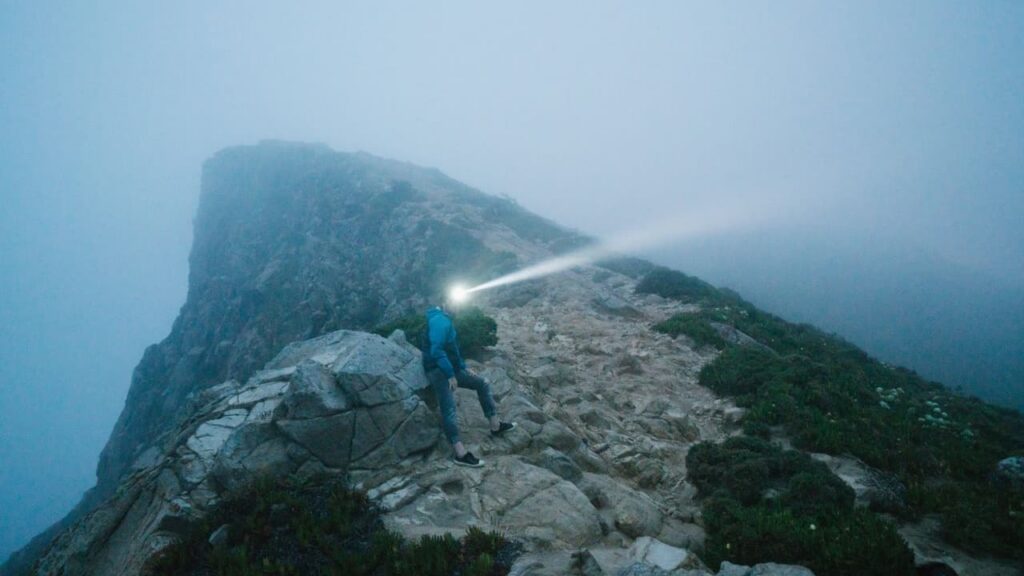 A hiker on a ridge trail at night with a beam of headlamp light on the path ahead, hands empty and hands free, the backcountry headlamp invisible against the dark