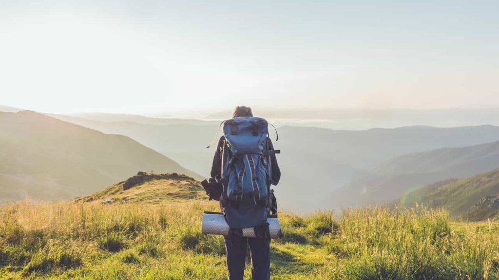 Hiker on a ridgeline with a water tube over the shoulder, sipping while walking, water bottle absent, pack sitting flush against the back, the backcountry hydration bladder made invisible by its use