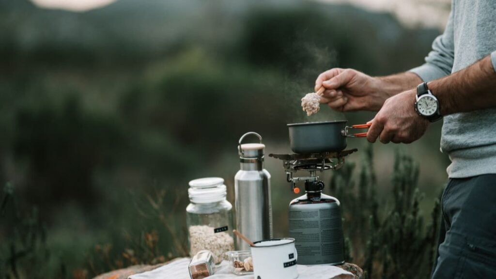 A small titanium canister stove simmering a single pot of tea on a rocky alpine ledge at dusk, a hiker's hands wrapped around a titanium cup, steam rising into cool evening air