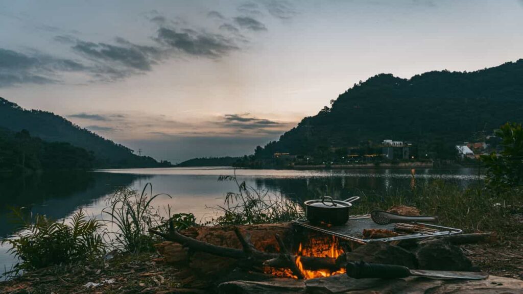 A minimalist titanium cookpot and pan on a campfire set beside a mountain stream, morning mist on the water, the cookware small and unobtrusive against the landscape