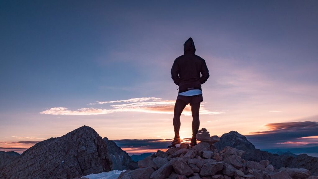 A hiker standing still on a rocky ridge at dawn, facing away from the camera, the mountain landscape stretching into mist, no pack visible, the body returned to the uncarved state of Pu
